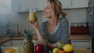 A woman drinking a smoothie in the kitchen with fruit on the counter.